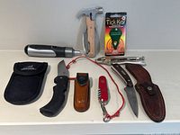 Full lot display of all hunting and survival tools including knives, multi-tool, pliers, tick removal tool, and sheaths on white surface