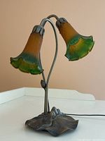 Front left angle of the lamp showing two stained glass lily-shaped shades in amber to green hues, mounted on metal curved stems and a lily pad shaped metal base.