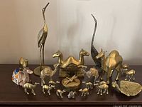 Photo showing the entire collection of brass animal figurines arranged on a dark surface. Includes two tall cranes, several camels of different sizes, and the trinket box.