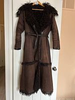 Full front view of long brown suede shearling coat hanging on hanger, showing oversized plush sheepskin collar, leather tie waist belt, and detailing along sleeves and hem.