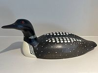 Side view of carved wooden loon decoy showing painted white spots and stripes on black body with red eye.