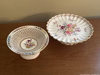 Both porcelain pedestal dishes side by side on a wooden surface; Dresden dish showing reticulated edge and colorful floral painting; Crown Staffordshire dish with scalloped gold trim and central floral design.