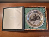 Opened presentation box showing sterling silver plate nestled in green satin lining alongside paperwork.