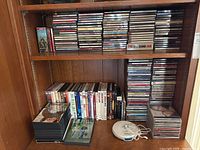 Shelf with large quantity of music CDs stacked alongside DVDs and a white Panasonic portable CD player with earphones