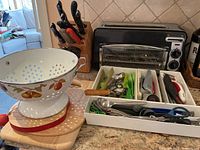 Wide view of slim black Hamilton Beach toaster oven with wooden Henkel knife block, white colander, wooden cutting board, bamboo trivet, and white tray with assorted utensils.