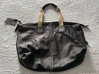 Front view of chocolate brown textured leather Roots bag with beige canvas handles and a black leather embossed logo patch at center.