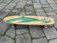 Side view of wooden Nash skateboard shaped like a small surfboard with green shark graphic and metal wheels on cobblestone ground.