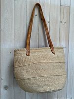 Full view of the woven sisal tote bag showing natural fiber body and brown leather straps hanging against light wooden wall.