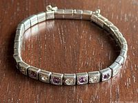 Full bracelet laid flat in a circular form on wooden surface, showing alternating purple and clear stones in square settings, with one clear stone missing.