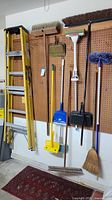 Photo showing a 6-foot yellow metal step ladder hanging on a pegboard wall with multiple cleaning brooms and tools around it.