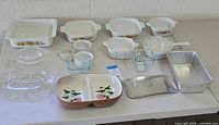 Full view of the assortment of vintage baking and serving dishes including Corning Ware Spice of Life and Cornflower Blue patterns, divided serving dish, aluminum loaf pan, and glass trays on a white table.