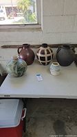 Five pottery vases placed on a white table in front of a window. Various shapes and colors including green, brown, checked tan and black.