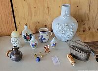 Overview of all items including large white ceramic vase with floral relief, smaller ceramic vases and pitchers, miniature decorative items, small copper pitcher, and wire wine bottle holder.