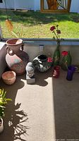 Photo showing large terracotta vase with handles and painted floral design, smaller pots including marbled black and white pot, small pinkish pot, stone Buddha head garden figure, green and red glass vases with flowers.