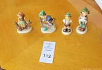 Lot of 4 vintage Hummel porcelain figurines displayed on a wooden table showing full lot and handwritten label card with lot number and information.
