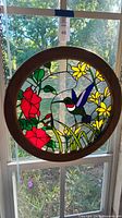 Front view of round stained glass panel showing hummingbird, flowers, and wooden frame hung in window.