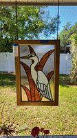 Frontal view of stained glass panel showing white bird with orange leaves framed in wood, hanging by chains outdoors.