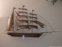 Front angle of large brass schooner wall art hanging on a textured beige wall with a small price tag attached near the hull.