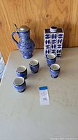 Full view of set showing cobalt blue ceramic beer stein, five cups, and vase on white table against wooden backdrop