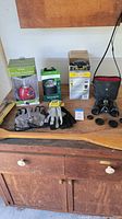 Lot items arranged on wood surface including boxed lanterns, work gloves, black binoculars with covers and case, and wooden paddle.