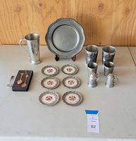 Full lot showing pewter plate with stand, four decorative pewter cups (tankard style), one taller pewter cup, six ceramic and pewter coasters, and three decorative spoons (two in a black display case).