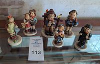 Photo showing all 6 Hummel figurines arranged in a row on glass shelf with auction number card visible.