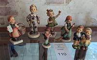 Six vintage Hummel figurines arranged on glass table surface with rustic background, displaying varying child characters and activities.