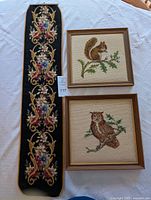 View of three needlepoint wall hangings: two framed squares featuring an owl and squirrel, one long narrow piece with floral design.