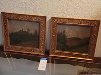Two landscape paintings in matching ornate gold tone frames, showing forested hills and cloudy skies