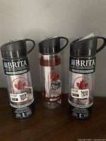 Photo of three Brita branded items: two water filter cartridges sealed in plastic packaging and one clear water bottle with black lid and handle, placed on a wooden surface.