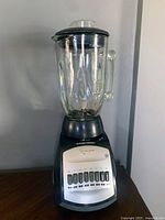 Full front view of Black & Decker blender on a counter against a gray wall, showing glass jar, black base, white control panel with push buttons, and black lid.