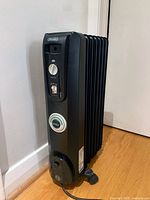 Photo of black DeLonghi oil-based heater positioned against wall on hardwood floor, showing front side with thermostat knob, power switch, and power cord.