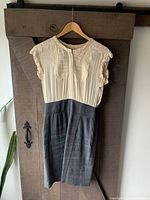 Full view of the Rebecca Taylor dress hanging on a wooden hanger against a barn wood style door. Dress has a cream ruched top with ruffled sleeves and dark gray skirt.