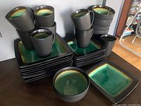 Photo showing stacks of matching square plates, bowls and mugs with distinctive green and brown interior glazing and matte black exterior.