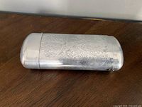 Closed silver-tone metal glasses case with engraved floral and scroll patterns, lying on wood surface.