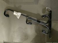 Photo of a black metal wall-mounted plant hanger bracket with curled hooks lying flat on a surface