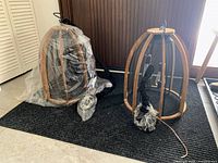 Two hanging cage light fixtures placed on the floor, one inside plastic wrap, both with wooden curved frames and black mesh cages.