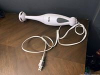 White Oster immersion blender with power cord coiled beside it, resting on a dark wood surface against a dark wall.