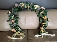 Full view of hand-made faux flower wreath with yellow roses, mixed flowers, green leaves, and light ribbons, placed on a beige couch.