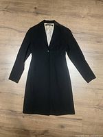 Full length view of the black long coat laid flat on a wooden surface showing the overall shape and single button closure.