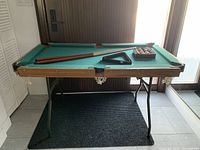 Front view of mini pool table with legs, green felt, cues, triangle rack, and pool balls.