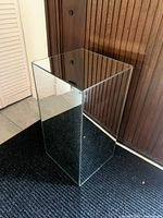 Photo of rectangular mirrored pedestal table with visible reflective surfaces and standing on dark carpet in front of wooden panel wall.