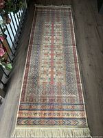 Photo of full length of thin decorative runner rug with geometric patterns and fringed edge, laid flat on wooden floor next to railing with flowers.