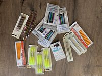 Photo showing various school supplies on a wooden surface including packaged and loose wood pencils, packs of black and blue ink pens, yellow highlighters, and mechanical pencil leads.