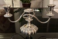 Silver-plated two-candle candelabra on a reflective black surface with floral patterns and a central handle for carrying, shown from the side angle.