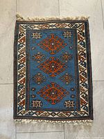Photo of a small rectangular rug with blue background and orange geometric designs and a white and brown border, taken on a tiled floor.