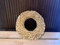 Photo of a circular wreath made from tightly arranged straw placed on a wooden surface against a vertical dark wood panel background.