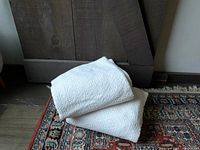 Two white throws with textured geometric patterns folded on a colorful patterned woven rug in front of a wooden background.