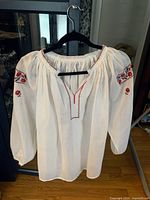 Front view of traditional Ukrainian blouse hung on black hanger showing white fabric with red and black floral embroidery on shoulders and neckline.