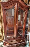 Front view of antique style mahogany curio cabinet showing arched top, glazed doors with carved floral accents, internal shelves, and bottom drawer with brass handles.